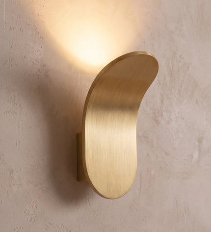 Vela wall light close-up: curved brushed gold plate with soft up light