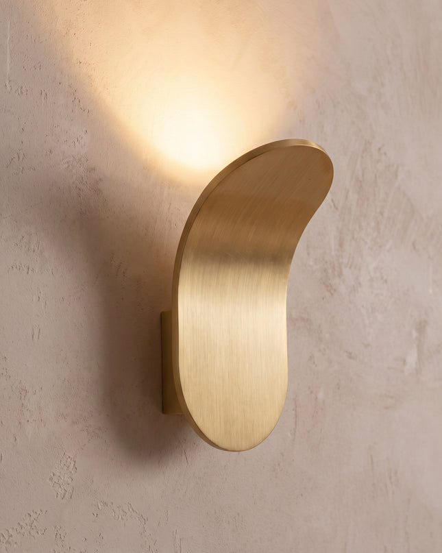 Vela wall light close-up: curved brushed gold plate with soft up light
